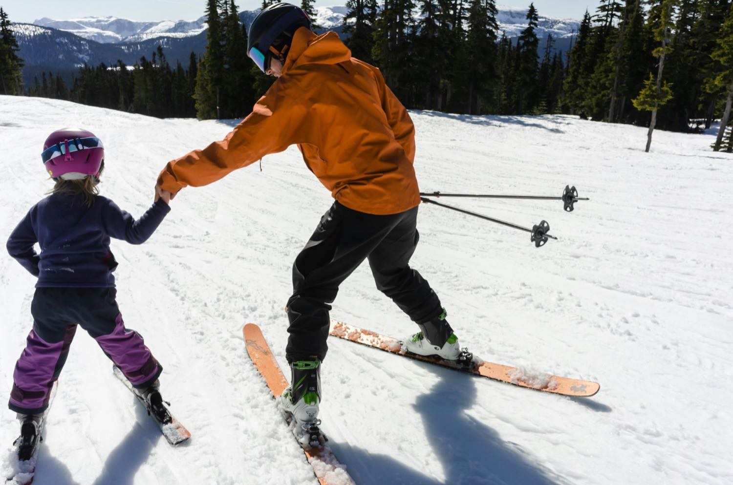 An adult holding a small child’s hand as she moves across a gentle snowy slope